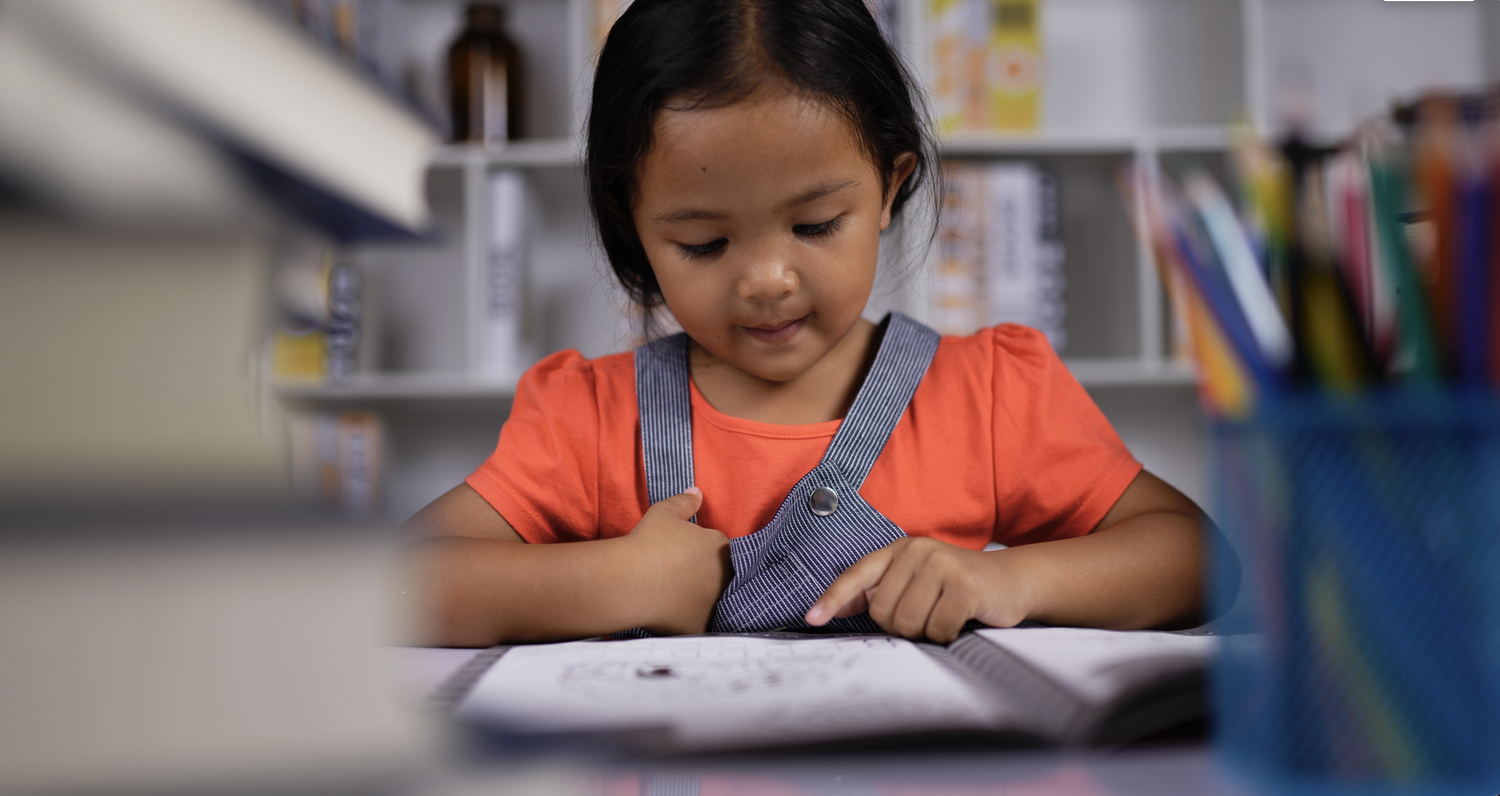 A young girl seated at a wooden table turning the pages of a large bilingual children's book